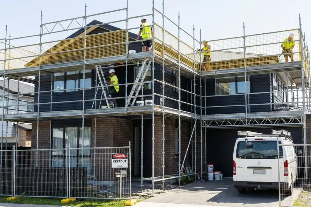 Construction workers on scaffolding performing exterior work on a new two-story residential house with dark siding and brick, with a van parked in the driveway.